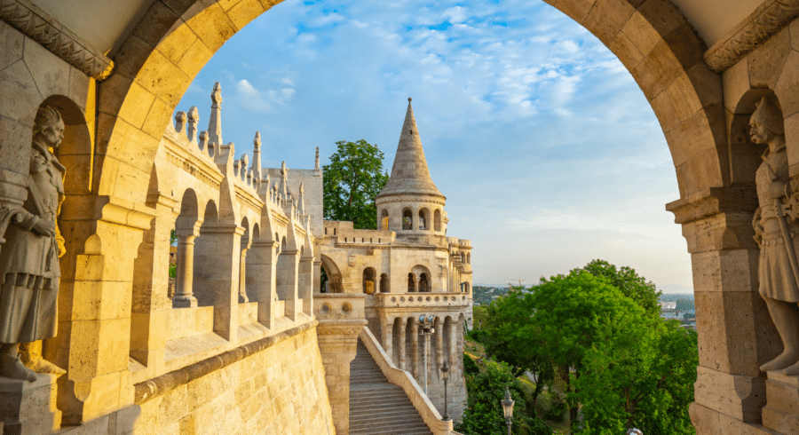 Stone archway frames the Fisherman’s Bastion, featuring turrets and spires with sculptured figures, under a partly cloudy sky with green trees in the background.