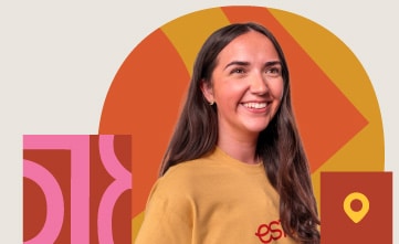 A woman with long brown hair, wearing a yellow shirt, stands smiling in front of geometric shapes in orange, yellow, and pink tones.
