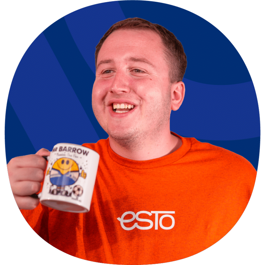 A smiling person in an orange Esto shirt holds a coffee mug with a Barrow football design against a blue background.