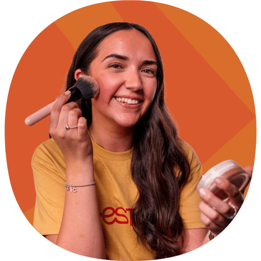 A woman with long brown hair smiles whilst applying make-up with a brush against an orange geometric background.