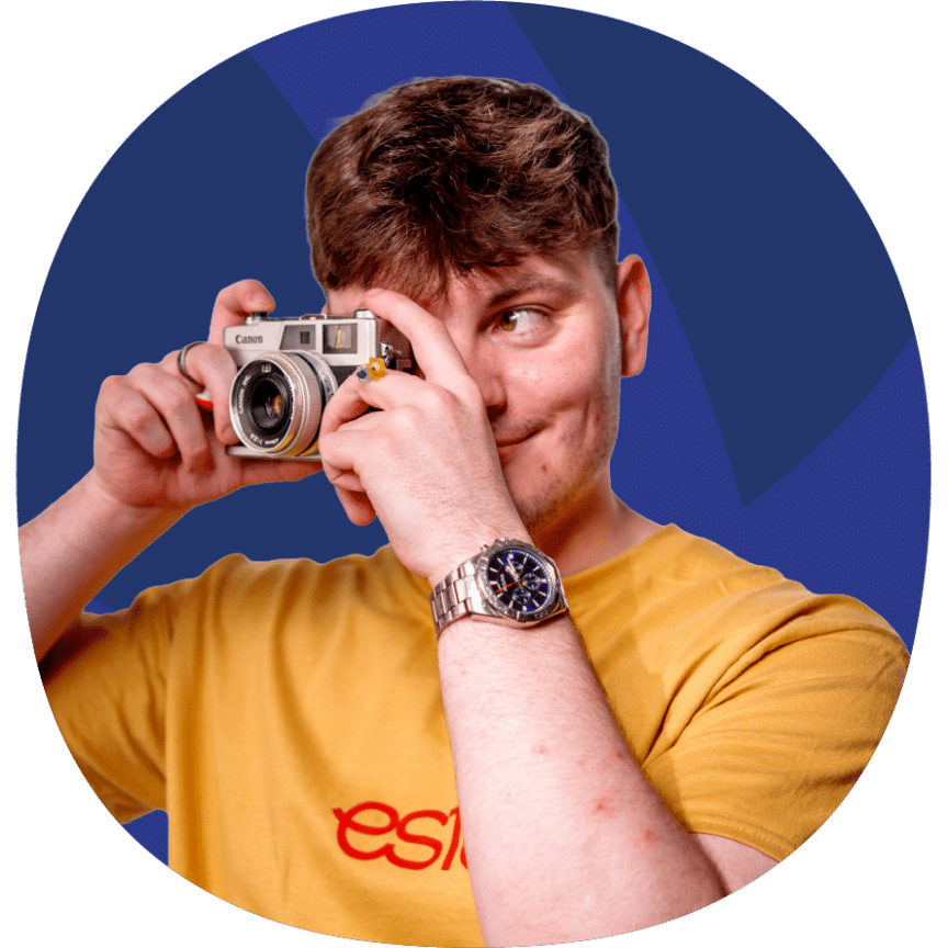 A person wearing a yellow shirt and wristwatch holds a vintage camera up to their face, posing against a blue background.