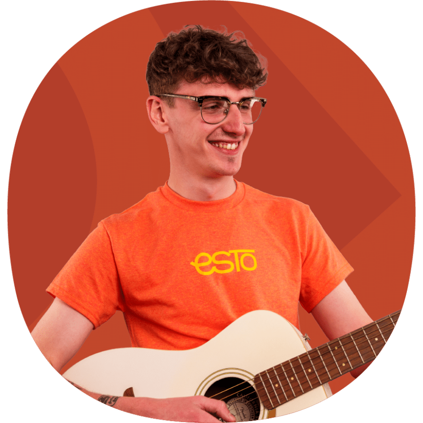 A young man wearing glasses and an orange "esto" t-shirt smiles whilst holding a white acoustic guitar against an orange background.