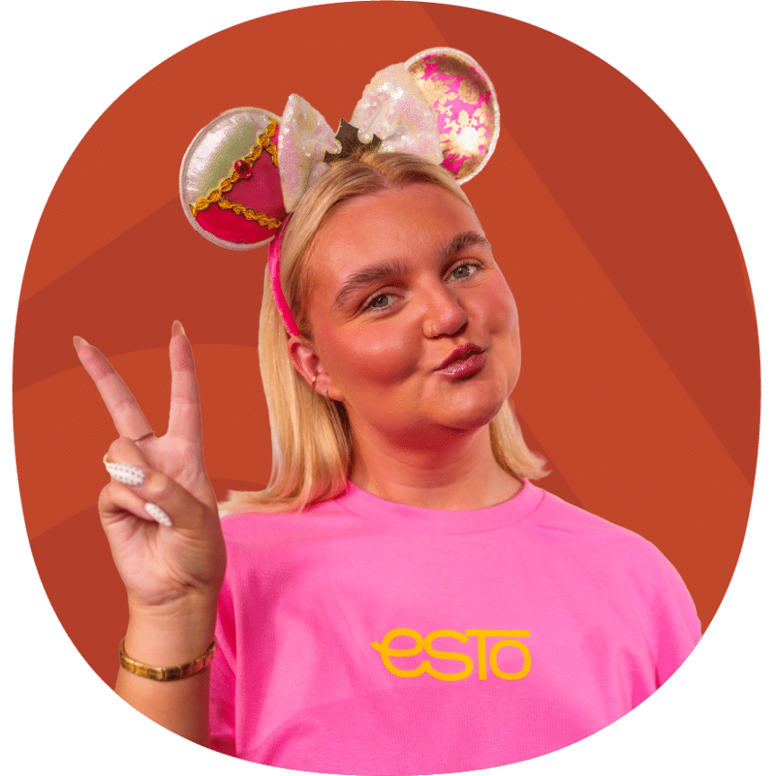 Woman wearing mouse ears and a bright pink shirt with "ESTO" text, making a peace sign with her hand, posing in front of an orange background.