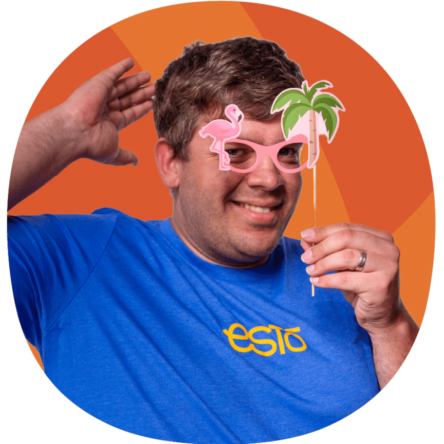 Man wearing a blue ESTÓ shirt poses with pink flamingo glasses and a palm tree prop in front of an orange background.
