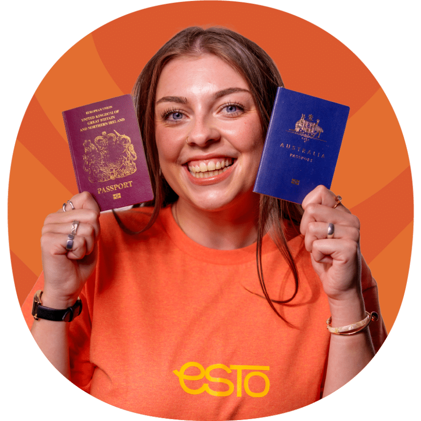 A woman in an orange shirt holds up a British passport in her left hand and an Australian passport in her right hand, smiling at the camera.