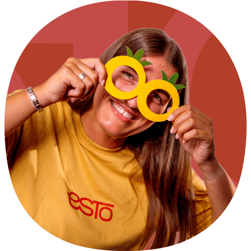 Smiling person in a yellow jumper holding up pineapple-shaped paper glasses in front of their eyes against a red background.