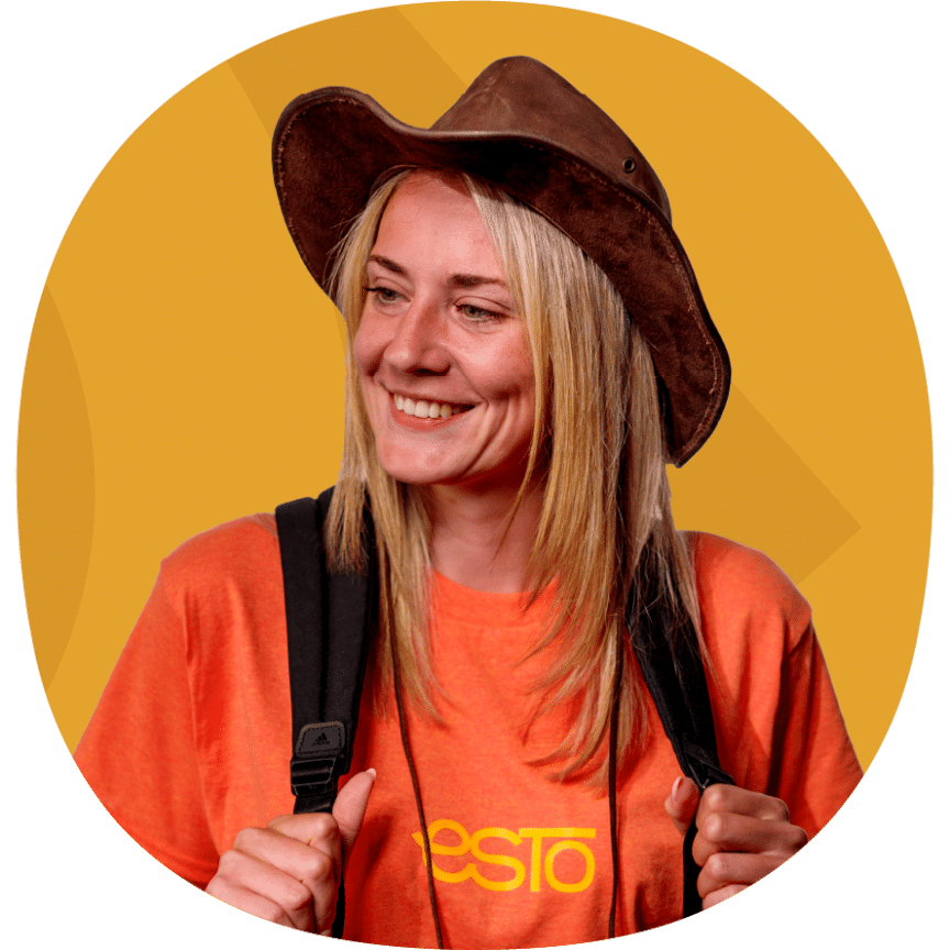 Smiling woman wearing a brown hat and orange T-shirt with "PESTO" text, carrying a black rucksack, standing in front of a yellow background.