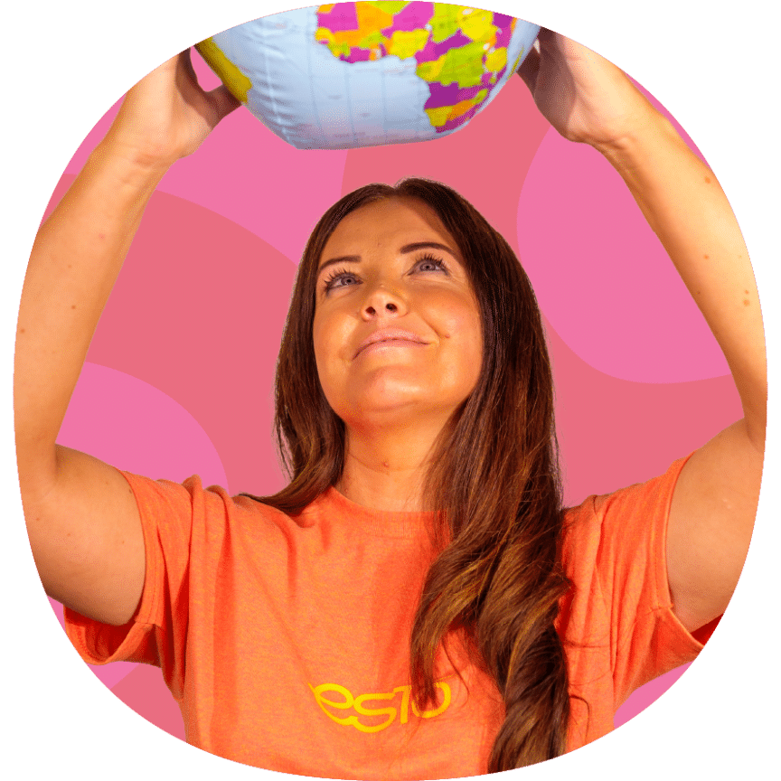 A woman in an orange shirt holds up an inflatable globe, looking up at it, against a pink background.