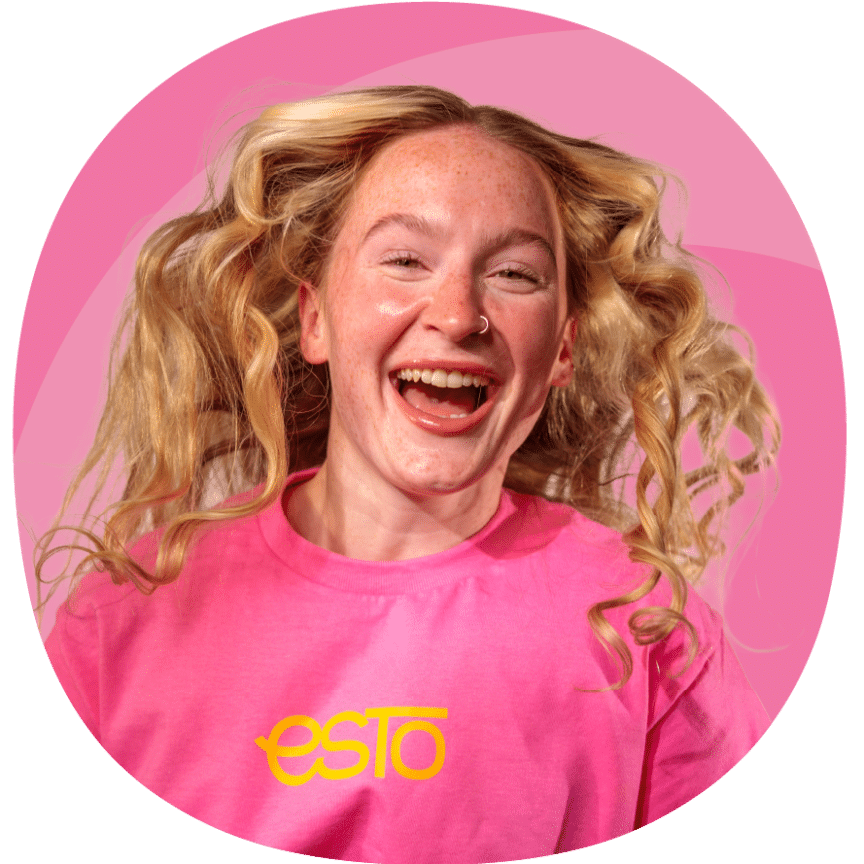 A person with curly blonde hair and freckles smiles widely whilst wearing a bright pink "esto" T-shirt against a pink background.