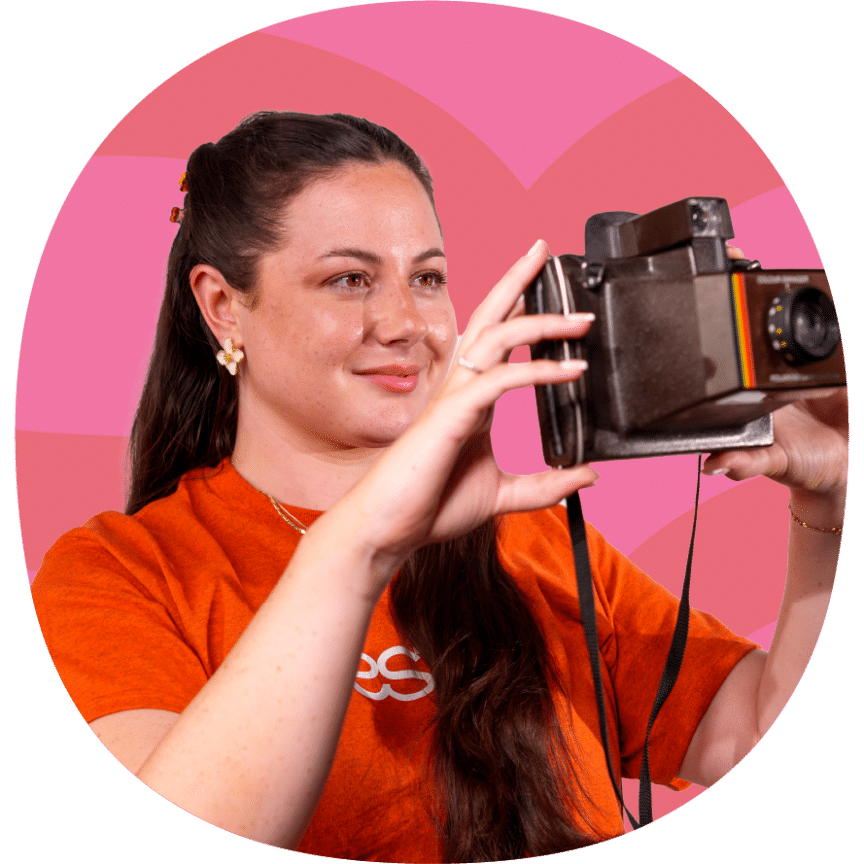 Woman in an orange shirt holding a vintage camera, standing in front of a pink patterned background.