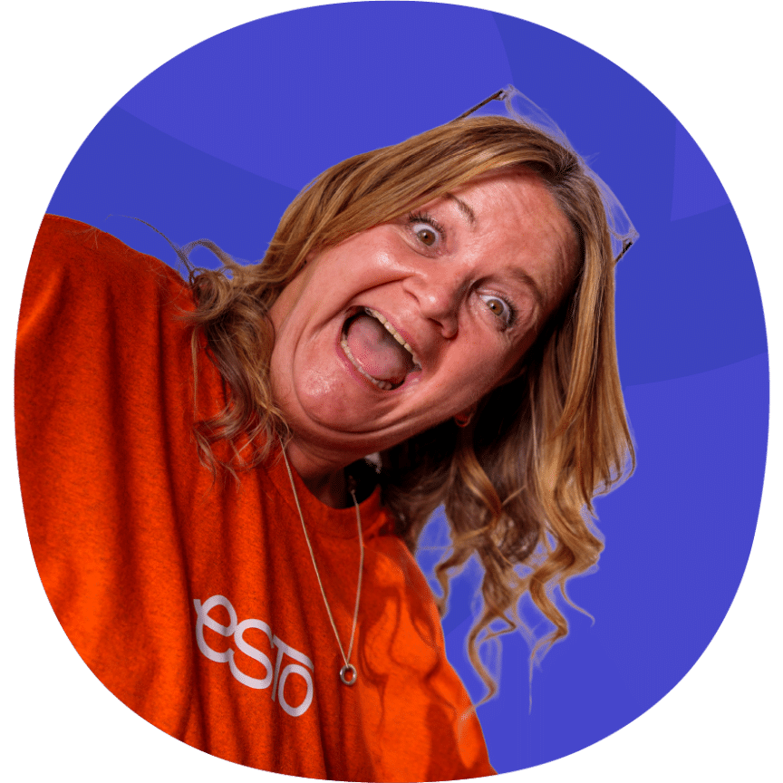 A woman with wavy hair wearing an orange shirt smiles widely and leans towards the camera against a purple background.