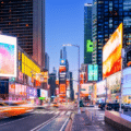 Times Square in New York City at dusk, featuring bright electronic billboards, city buildings, and blurred lights from moving traffic.