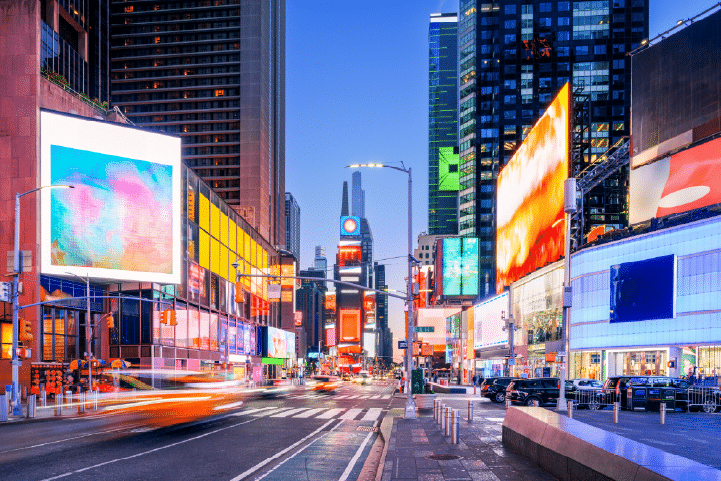 Times Square in New York City at dusk, featuring bright electronic billboards, city buildings, and blurred lights from moving traffic.