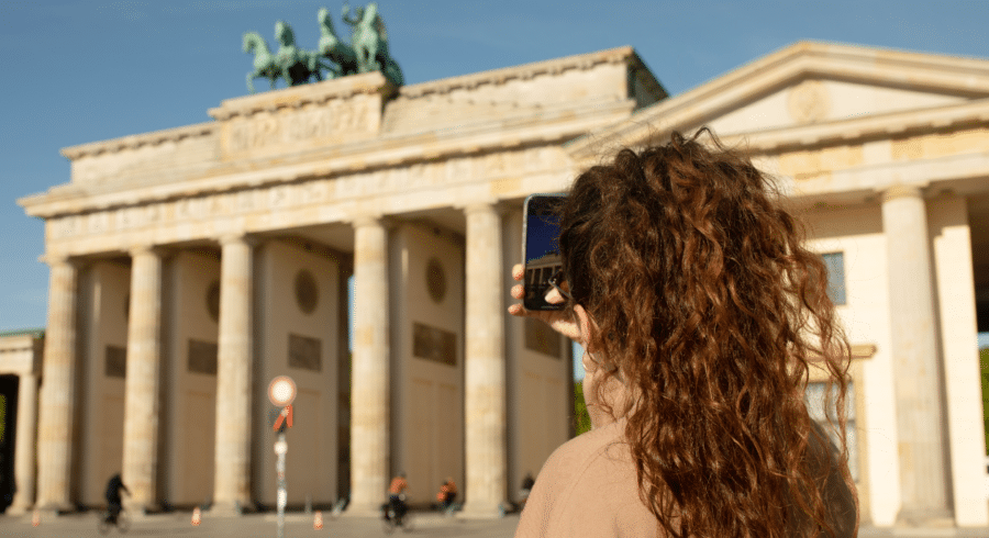 A person with curly hair takes a photo of the Brandenburg Gate in Berlin, Germany, on a sunny day.