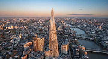 Aerial view of The Shard skyscraper in London, surrounded by city buildings, with the River Thames visible in the background at sunset.