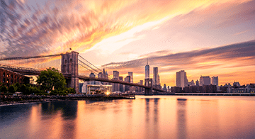 The Brooklyn Bridge spans the East River with the New York City skyline in the background at sunset, reflected in the calm water.