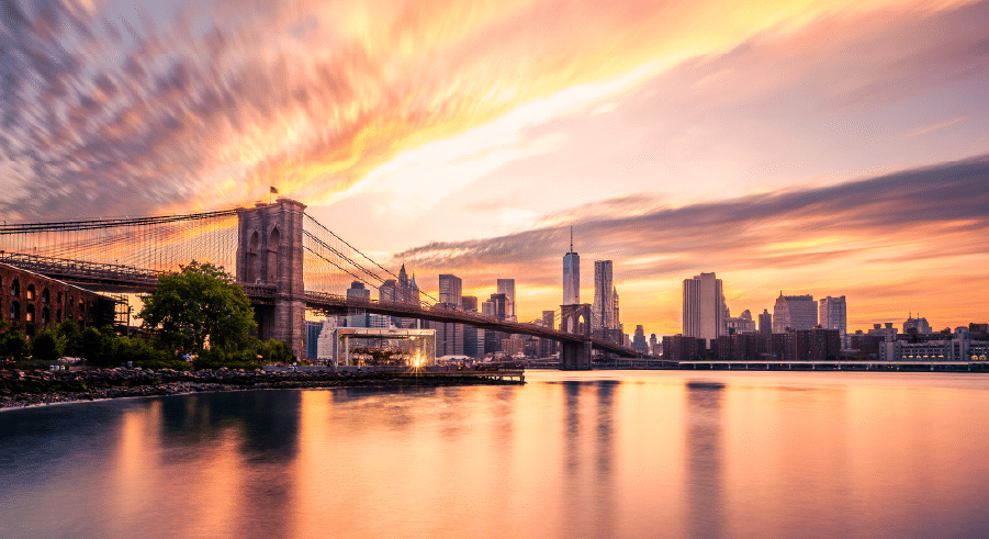 The Brooklyn Bridge spans the East River at sunset, with the New York City skyline and dramatic clouds in the background, reflected in the water.