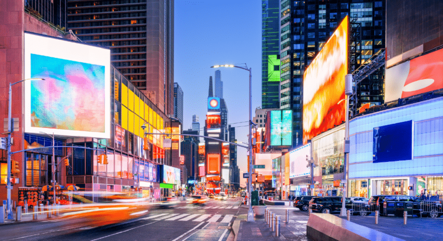 Times Square in New York City at dusk, with bright digital billboards, tall buildings, light trails from cars, and a mostly empty street.