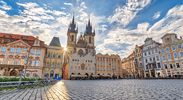 A historic town square with cobblestone paving, colourful buildings, and a large Gothic church with two tall spires under a partly cloudy sky.