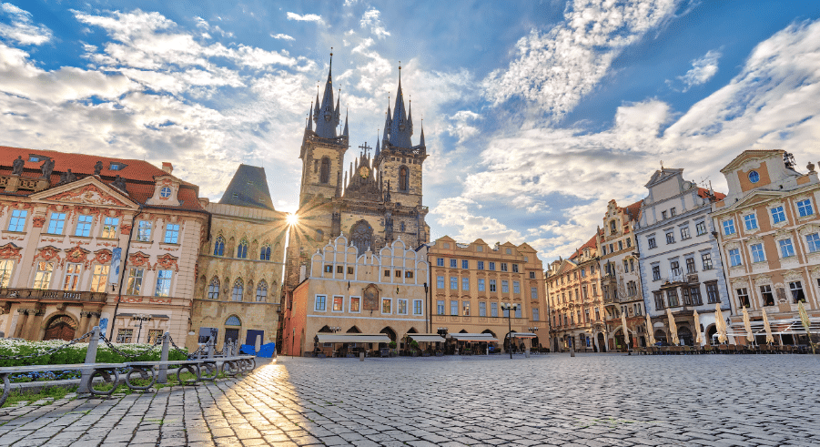 A cobblestone square with historic buildings and a large Gothic church with two tall spires under a partly cloudy sky.