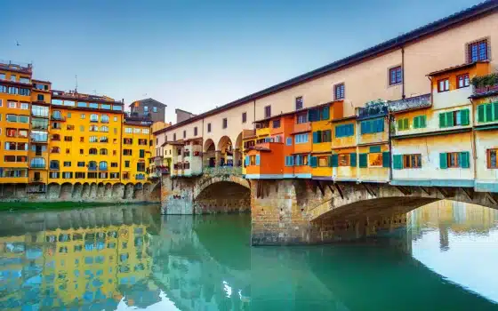 Colourful buildings and shops line the historic Ponte Vecchio bridge over the Arno River in Florence, Italy, under a clear sky.