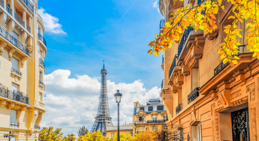 Eiffel Tower in the background between classic Parisian buildings, with blue sky and autumn trees in the foreground.
