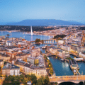 Aerial view of Geneva, Switzerland, showing the Jet d’Eau fountain on Lake Geneva, city buildings, and surrounding mountains at dusk.
