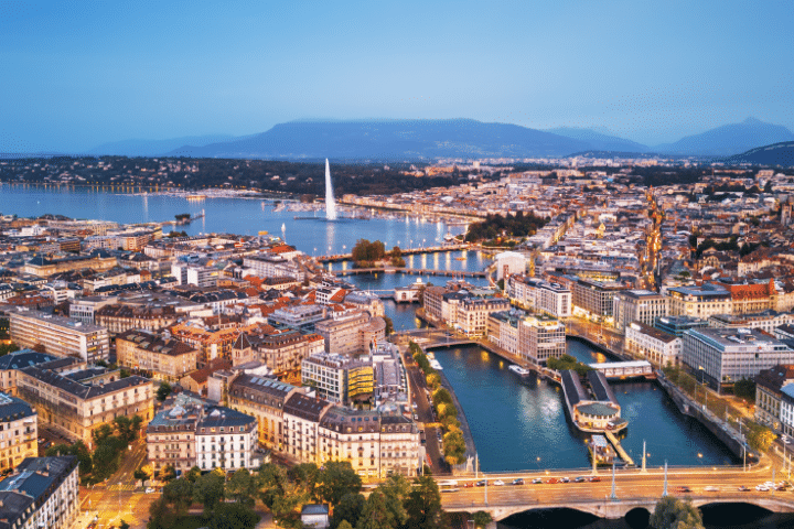 Aerial view of Geneva at dusk, showing the Jet d’Eau fountain, the Rhône River, and city buildings with mountains in the background.
