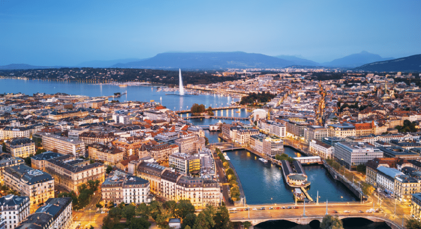 Aerial view of Geneva at dusk, showing the Jet d’Eau fountain, the Rhône River, and city buildings with mountains in the background.