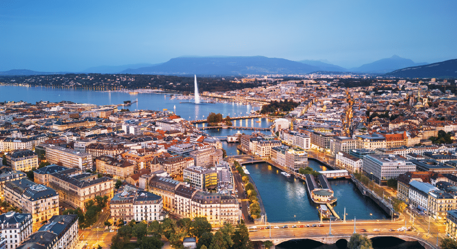 Aerial view of Geneva at dusk, showing the Jet d’Eau fountain, the Rhône River, and city buildings with mountains in the background.