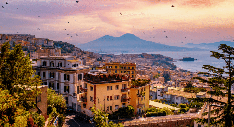 Cityscape of Naples, Italy at sunset with Mount Vesuvius in the background, buildings in the foreground, and birds flying in the sky.