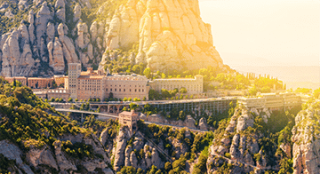 A large monastery complex is built into a rocky mountainside, surrounded by steep cliffs and greenery, under bright sunlight.