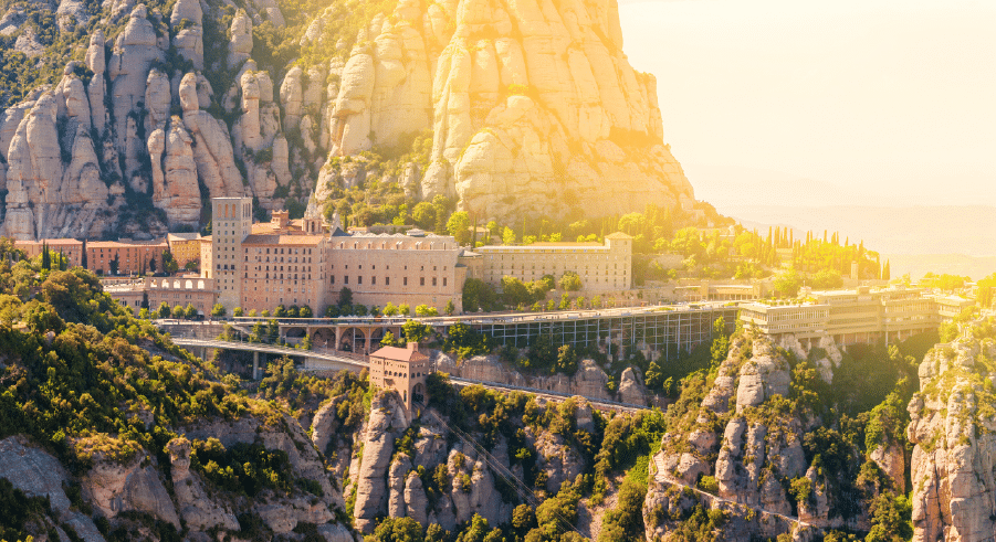 A large monastery complex is nestled among rocky mountains with sunlight shining over the landscape.