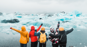 Five people in colourful jackets stand with arms raised, facing a landscape of melting icebergs and water under a cloudy sky.