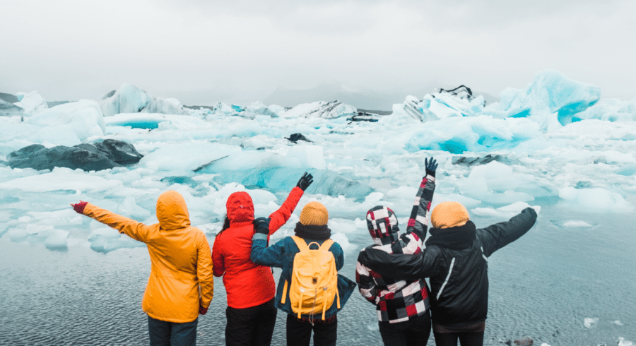 Five people in colourful jackets stand with arms raised, facing a landscape of icebergs and water under a cloudy sky.