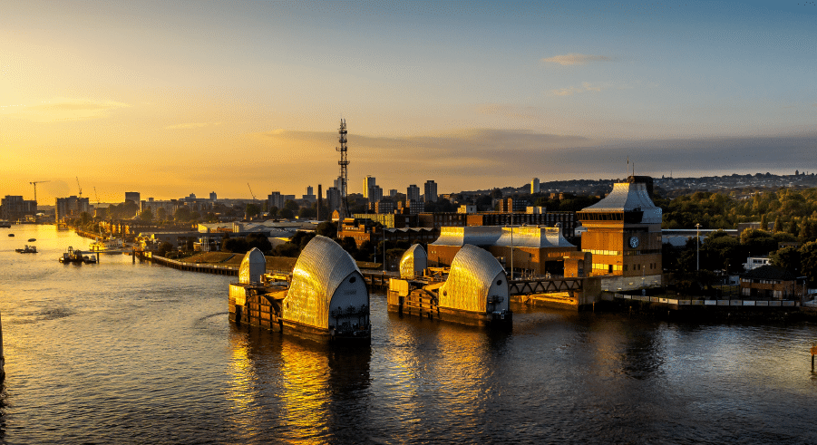 The Thames Barrier on the River Thames at sunset, with the London skyline and buildings visible in the background.
