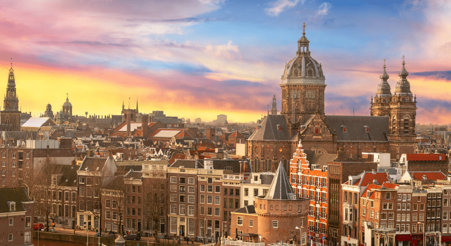 Cityscape of Amsterdam at sunset, featuring historic buildings, church spires, and a colourful sky in the background.