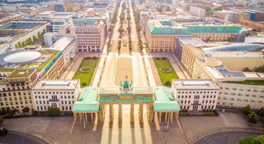 Aerial view of the Brandenburg Gate in Berlin, Germany, with adjacent historic buildings and a wide, tree-lined boulevard extending into the distance.