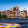 The Reichstag building in Berlin at dusk, with its glass dome illuminated and a blurred boat passing on the Spree river in the foreground.
