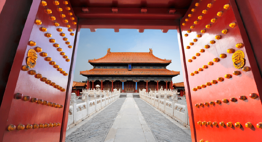 View through large red doors with golden studs towards a traditional Chinese palace building in the Forbidden City, Beijing, featuring ornate architecture and stone pathways.