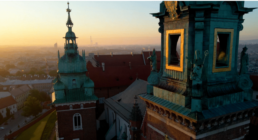 Aerial view of historic church towers with green copper roofs at sunset, overlooking a cityscape with buildings and a hazy sky in the background.