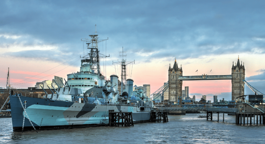 A large historic warship is moored on the River Thames with London's Tower Bridge visible in the background at dusk.