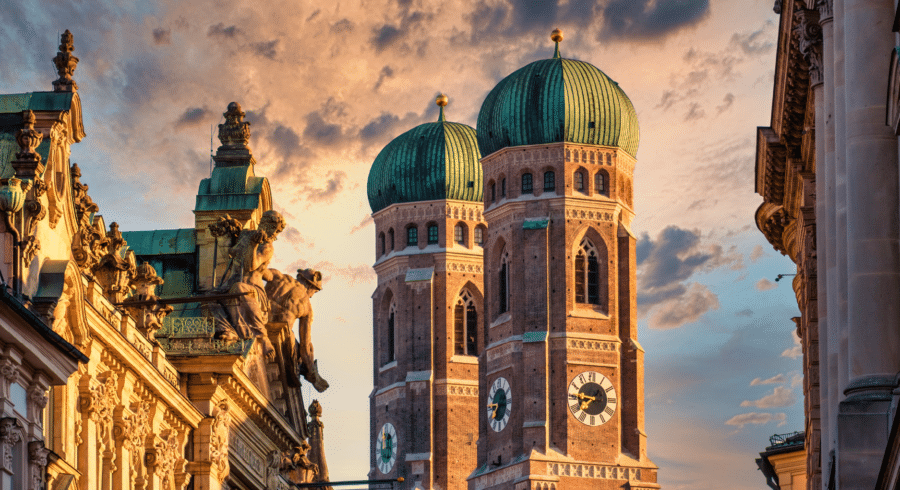 Two towers with green domes and clock faces rise above surrounding historic buildings under a cloudy sky at sundown.