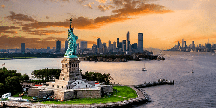 The Statue of Liberty stands on Liberty Island with the New York City skyline and a colourful sunset in the background.
