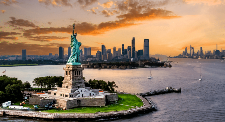 The Statue of Liberty stands on Liberty Island with the New York City skyline in the background at sunset, surrounded by water and a few boats.