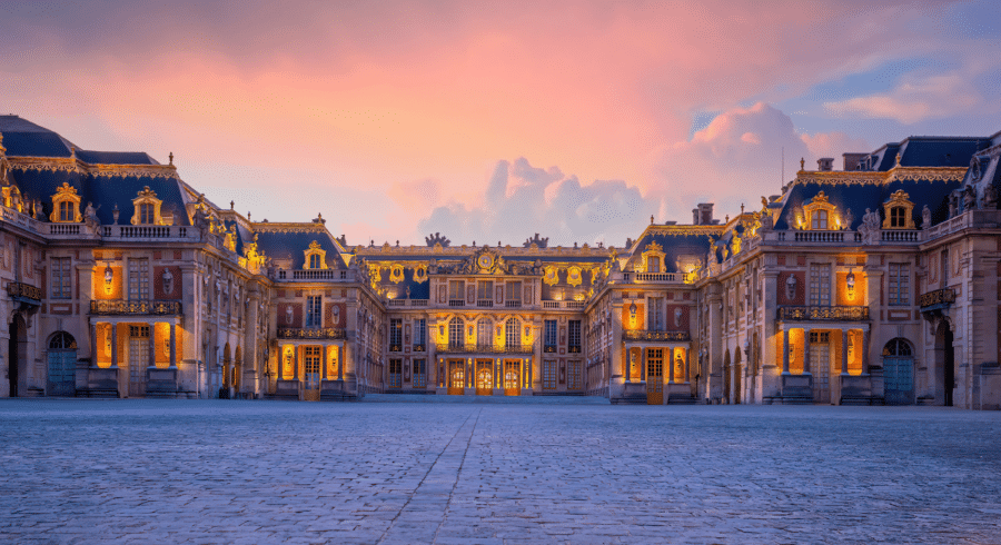 The illuminated façade of the Palace of Versailles is shown at dusk, with a wide cobbled courtyard in the foreground and a colourful sky in the background.
