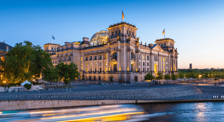 The Reichstag building in Berlin, Germany, with its glass dome and surrounding flags, is illuminated at dusk beside a river with a blurred boat passing in the foreground.