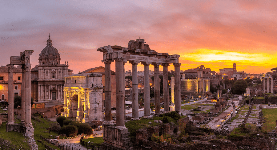 Ancient Roman ruins, including several tall columns and arches, stand at sunset in the Roman Forum in Rome, Italy, with historic buildings in the background.