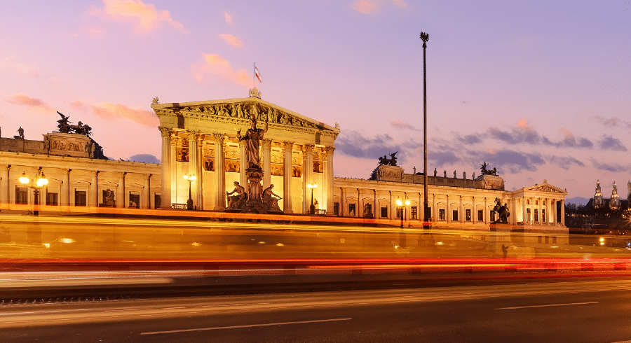 The illuminated Austrian Parliament Building in Vienna at dusk, with blurred lights from passing vehicles in the foreground.