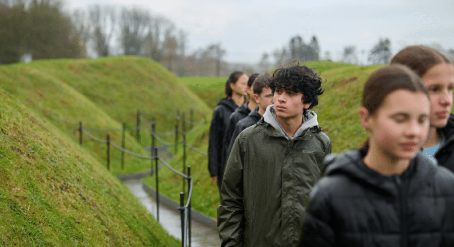 A group of young people walks along a narrow path bordered by grassy mounds on a cloudy day.